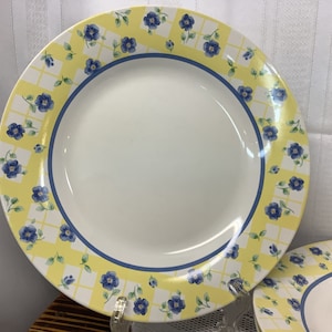 May include: White ceramic plate with a yellow and white checkered border. The border is decorated with blue floral designs and a blue trim. The plate is part of a dinnerware set.