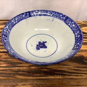 May include: A white ceramic bowl with a blue floral design around the rim and a blue floral motif in the center. The bowl is set on a wooden surface.