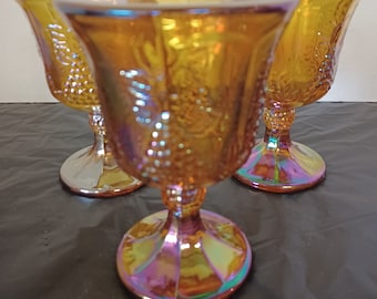 May include: Three iridescent amber-colored goblets with a grape design. The goblets have a flared rim, a detailed bowl, and a pedestal base. The glass reflects a rainbow of colors. The goblets are likely vintage or antique.