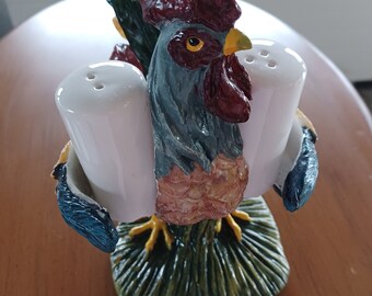 May include: A ceramic rooster-shaped salt and pepper shaker set. The rooster has a red comb, blue-gray feathers, and yellow feet. The salt and pepper shakers are white and held in place by the rooster's body. The base is green with a grass-like texture.