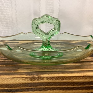 May include: A light green glass serving dish with a handle. The dish has a scalloped edge and a decorative handle with a circular opening. The dish is on a wooden surface.