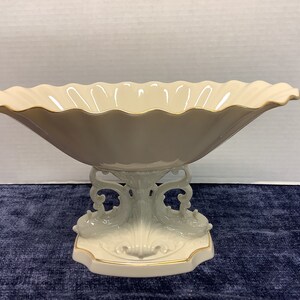 May include: A decorative, off-white ceramic centerpiece bowl with a scalloped rim and gold trim. The bowl is supported by a pedestal base featuring dolphin and scroll designs. A stylish addition to any home decor.