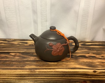 May include: A brown ceramic teapot with a rounded body, a curved handle, and a spout. The teapot features a reddish-brown floral design and is adorned with an orange string. The teapot is placed on a wooden surface.