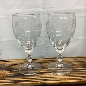May include: Two clear wine glasses with etched leaf designs. The glasses have a rounded bowl, a decorative stem, and a circular base. They are displayed on a wooden surface against a white backdrop.