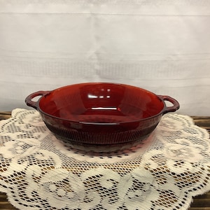 May include: A ruby red glass casserole dish with two handles. The dish has a ribbed design around the sides and is sitting on a cream-colored lace doily. The background is a white tablecloth.
