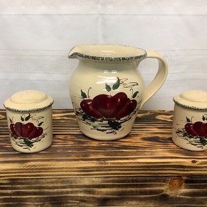 May include: A cream-colored ceramic pitcher and two salt and pepper shakers. Each piece is decorated with a red apple design and green leaves. The pitcher has a handle and spout. The set is displayed on a wooden surface.