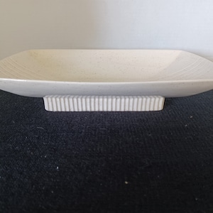 May include: A cream-colored, rectangular dish with a speckled surface, resting on a ribbed base. The dish has gently curved sides and subtle linear detailing. This decorative piece is suitable for serving food or as a display item.
