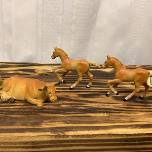 May include: A collection of miniature farm animal figurines, including two running foals and a resting bull. The figurines are painted in shades of tan and brown, with detailed features. The scene is set on a rustic wooden surface.
