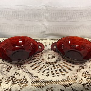 May include: Two ruby red glass bowls with handles. The bowls have a ribbed design and are sitting on a white lace doily. The bowls are likely vintage or antique and are suitable for serving.