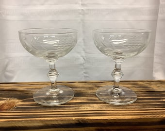 May include: Two clear, stemmed champagne glasses with etched leaf designs. The glasses have a wide, shallow bowl and a decorative stem. They are displayed on a wooden surface against a white background.