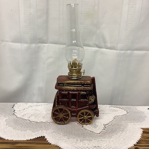 May include: A decorative oil lamp shaped like a stagecoach. The lamp features a clear glass chimney, a brass burner, and a red and gold painted base with spoked wheels. The lamp sits on a wooden surface.