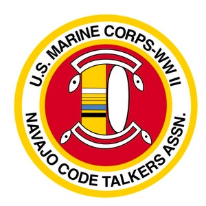 Op de afbeelding: Een geel en rood cirkelvormig embleem met de tekst "U.S. MARINE CORPS-WW II NAVAJO CODE TALKERS ASSN." rond de rand. Het midden van het embleem toont een gestileerde afbeelding van een symbool van een Navajo-code-spreker.