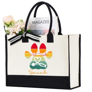 May include: A white canvas tote bag with black handles and a black bottom. The front features a colorful paw print design with the words "Vet Tech" and a stethoscope. The name "Sarah" is written below the paw print. The bag contains flowers and a magazine.