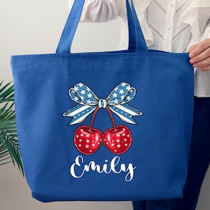 May include: Blue tote bag with a cherry design. The cherries are red with white star accents, and the bow is blue and white. The name "Emily" is written in white script below the cherries. The bag has blue handles.