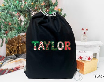 May include: Black Christmas gift bag with a personalized name "Taylor" in festive red, green, and white letters. The letters are decorated with Christmas patterns like candy canes, snowflakes, and plaid.