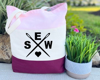 May include: A tri-color tote bag with a pink, white, and burgundy design. The bag has a white panel with the letters "S" and "W" on either side of a heart with the letter "E" above the heart. Two crossed needles are above the letter "E". The text "Tri-Color Pink" is below the bag.
