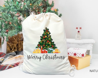 May include: A white canvas Christmas gift bag with a printed image of a Christmas tree with ornaments and houses. The text "Merry Christmas" is printed below the image. The bag is on a white surface.
