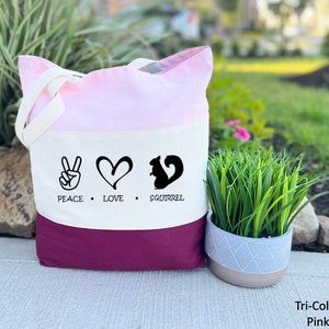 May include: A tri-color tote bag with a pink, white, and burgundy design. The bag has a peace sign, heart, and squirrel silhouette with the text "Peace Love Squirrel".
