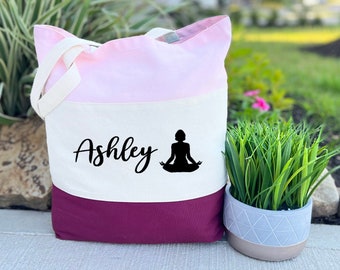 May include: A tri-color tote bag with pink, white, and burgundy fabric. The bag has a white panel with the name "Ashley" and a black silhouette of a person in a yoga pose.