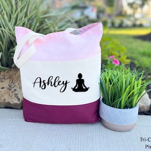 Pu&ograve; includere: Una borsa tote tricolore in tessuto rosa, bianco e bordeaux. La borsa ha un pannello bianco con il nome "Ashley" e una silhouette nera di una persona in una posa yoga.