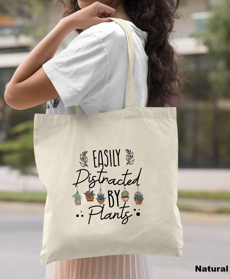 Pode incluir: Uma bolsa tote de lona natural com texto preto que diz "Easily Distracted By Plants" e apresenta um gr&aacute;fico de plantas em vasos pendurados no texto.
