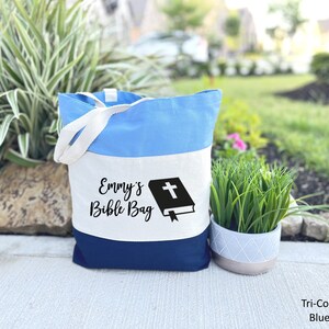 May include: A tri-color tote bag with a white stripe that says "Emmy's Bible Bag" and a black silhouette of a bible with a cross on the cover. The bag is blue and navy blue.