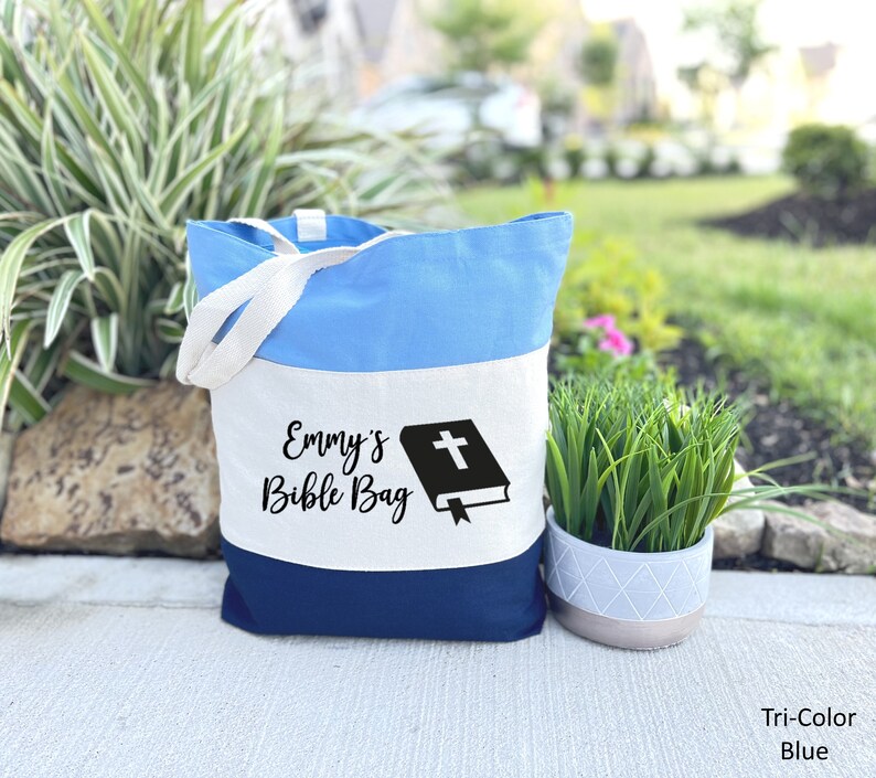 May include: A tri-color tote bag with a white stripe that says "Emmy's Bible Bag" and a black silhouette of a bible with a cross on the cover. The bag is blue and navy blue.