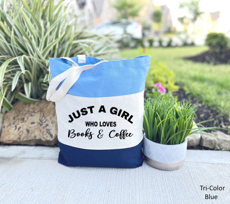 Puede incluir: Una bolsa de tela de color tricolor con tejido azul, blanco y azul marino. La bolsa tiene dos asas blancas y el texto "Just a girl who loves books & coffee" impreso en la secci&oacute;n de tela blanca.