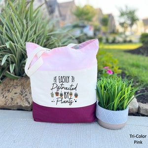 Pode incluir: Uma bolsa tote de lona branca com um design rosa e bord&ocirc;. A bolsa tem um gr&aacute;fico preto que diz "Easily Distracted by... Plants" com uma fileira de pequenas plantas em vasos penduradas no texto.