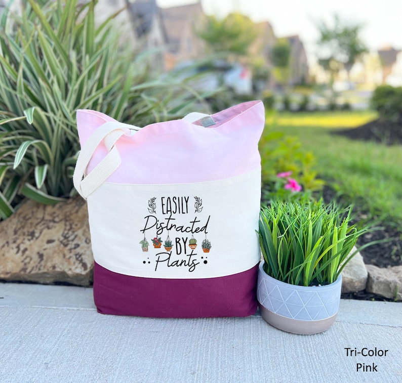 Pode incluir: Uma bolsa tote de lona branca com um design rosa e bord&ocirc;. A bolsa tem um gr&aacute;fico preto que diz "Easily Distracted by... Plants" com uma fileira de pequenas plantas em vasos penduradas no texto.