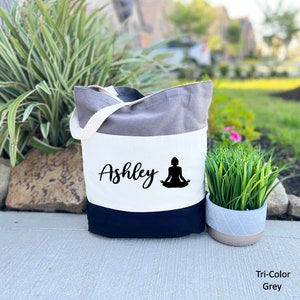 Pu&ograve; includere: Una borsa tote in tela grigia a tre colori con manici neri. La borsa ha un pannello bianco con il nome "Ashley" e una silhouette nera di una persona in una posa yoga.