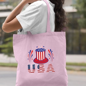 May include: A light pink tote bag with a red, white, and blue graphic of a top hat with fireworks and the word "USA" in a distressed American flag font.