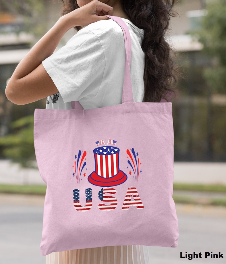 May include: A light pink tote bag with a red, white, and blue graphic of a top hat with fireworks and the word "USA" in a distressed American flag font.