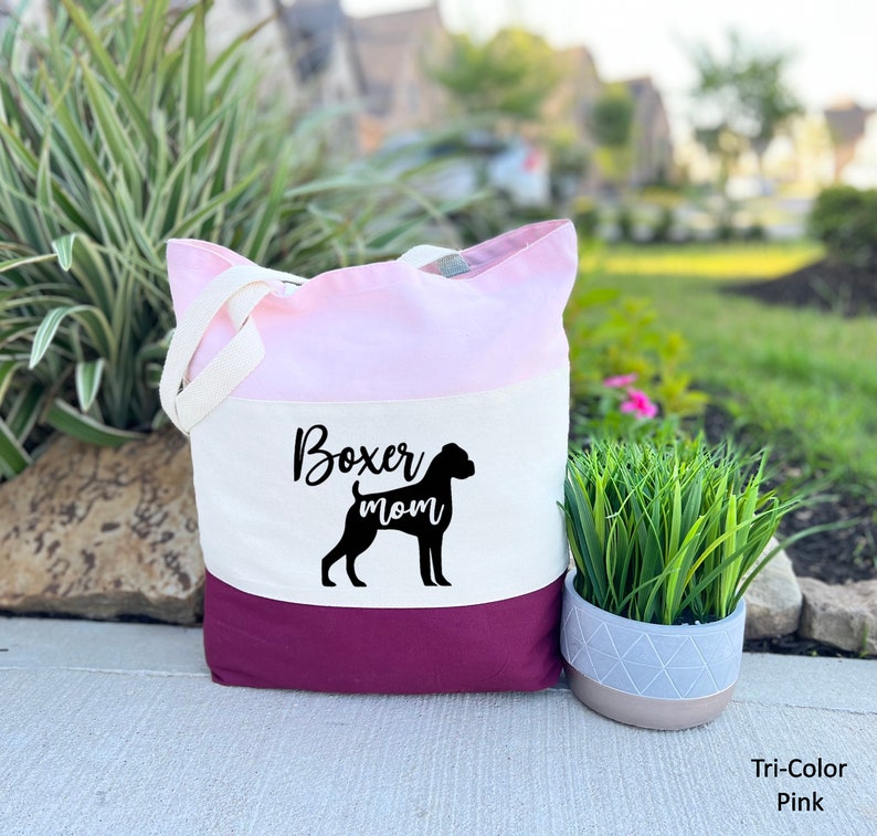 Puede incluir: Una bolsa de mano tricolor con un dise&ntilde;o rosa, blanco y burdeos. La bolsa tiene una silueta negra de un perro boxer y el texto "Boxer mom" impreso en la secci&oacute;n blanca.