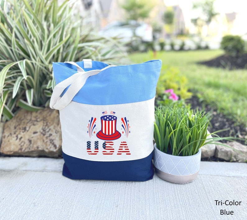 May include: A blue, white, and navy blue canvas tote bag with a red, white, and blue patriotic design. The design features a top hat with stars and stripes, fireworks, and the word "USA".