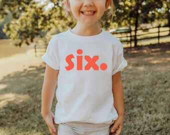 Può includere: Maglietta bianca con la parola "six" in lettere arancioni a blocchi. La maglietta è indossata da un bambino e lo sfondo è una scena sfocata all'aperto con alberi ed erba.