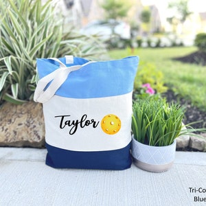 K&ouml;nnte beinhalten: Eine blaue, wei&szlig;e und marineblaue Canvas-Tragetasche mit dem Namen "Taylor" und einem gelben Pickleball, der auf der Vorderseite aufgedruckt ist.