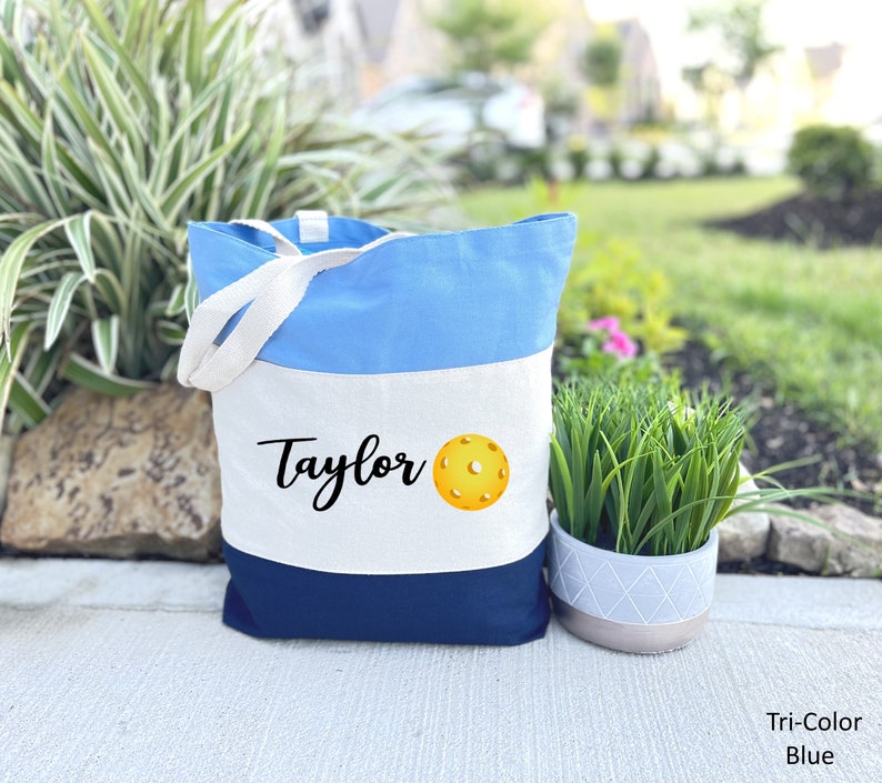 K&ouml;nnte beinhalten: Eine blaue, wei&szlig;e und marineblaue Canvas-Tragetasche mit dem Namen "Taylor" und einem gelben Pickleball, der auf der Vorderseite aufgedruckt ist.