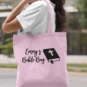 May include: A light pink tote bag with the text "Emmy's Bible Bag" and a black silhouette of a closed book with a cross on the cover.