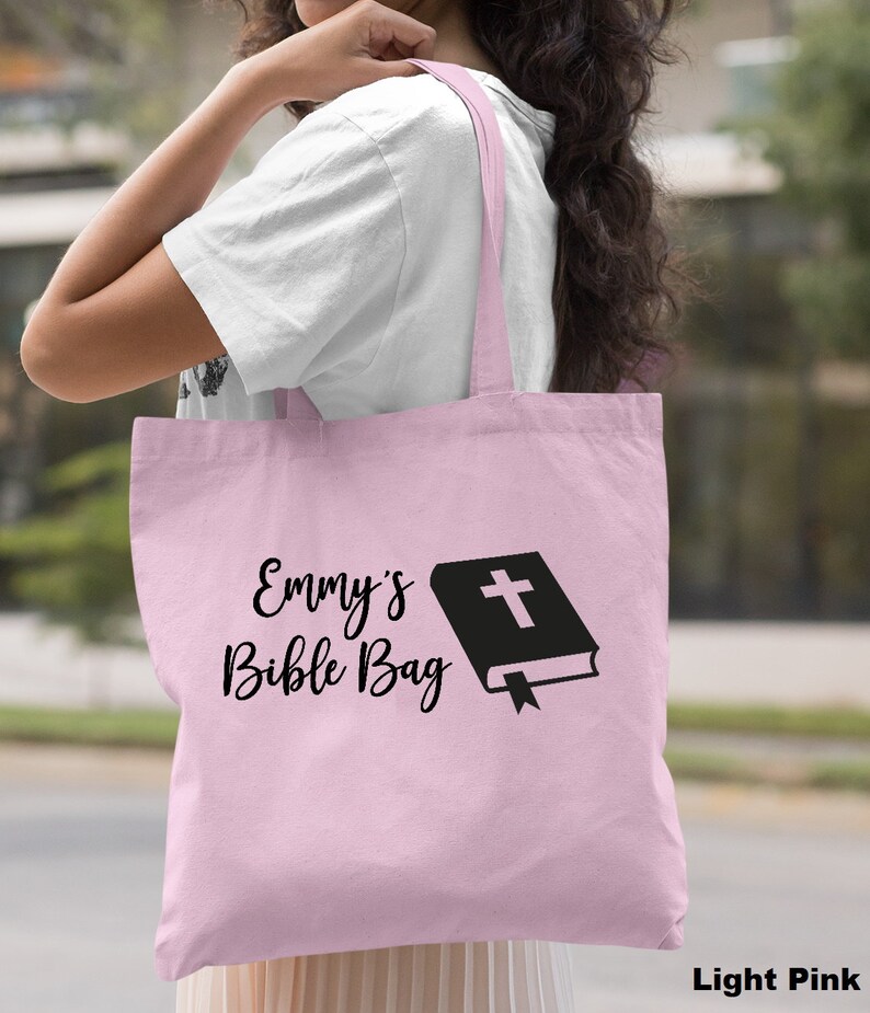May include: A light pink tote bag with the text "Emmy's Bible Bag" and a black silhouette of a closed book with a cross on the cover.