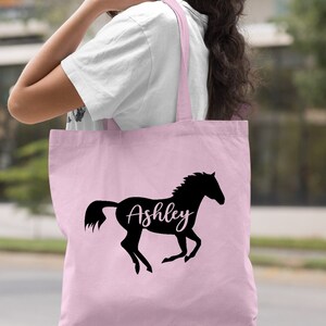May include: A light pink tote bag with a black silhouette of a horse and the name "Ashley" written in white inside the horse.