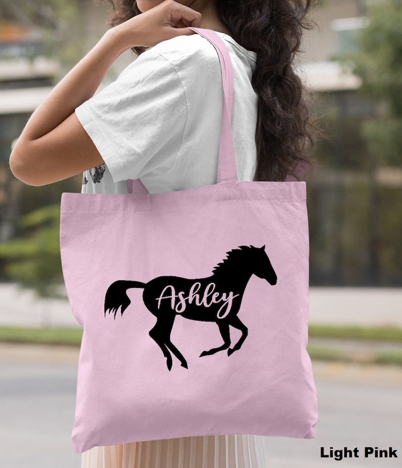 May include: A light pink tote bag with a black silhouette of a horse and the name "Ashley" written in white inside the horse.