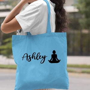 Pu&ograve; includere: Borsa tote blu chiaro con il nome "Ashley" e una silhouette di una persona in una posa di yoga.