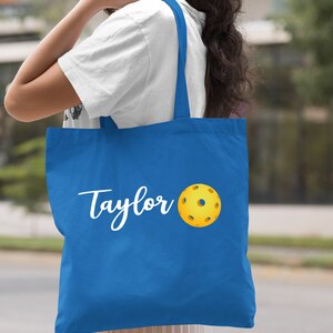 K&ouml;nnte beinhalten: Eine k&ouml;nigsblaue Tragetasche mit wei&szlig;em Text, der "Taylor" sagt, und einem gelben Pickleball.