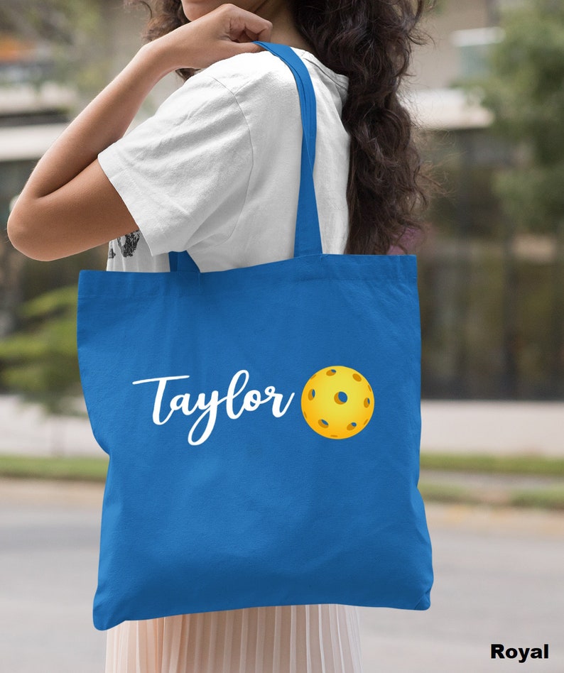 K&ouml;nnte beinhalten: Eine k&ouml;nigsblaue Tragetasche mit wei&szlig;em Text, der "Taylor" sagt, und einem gelben Pickleball.