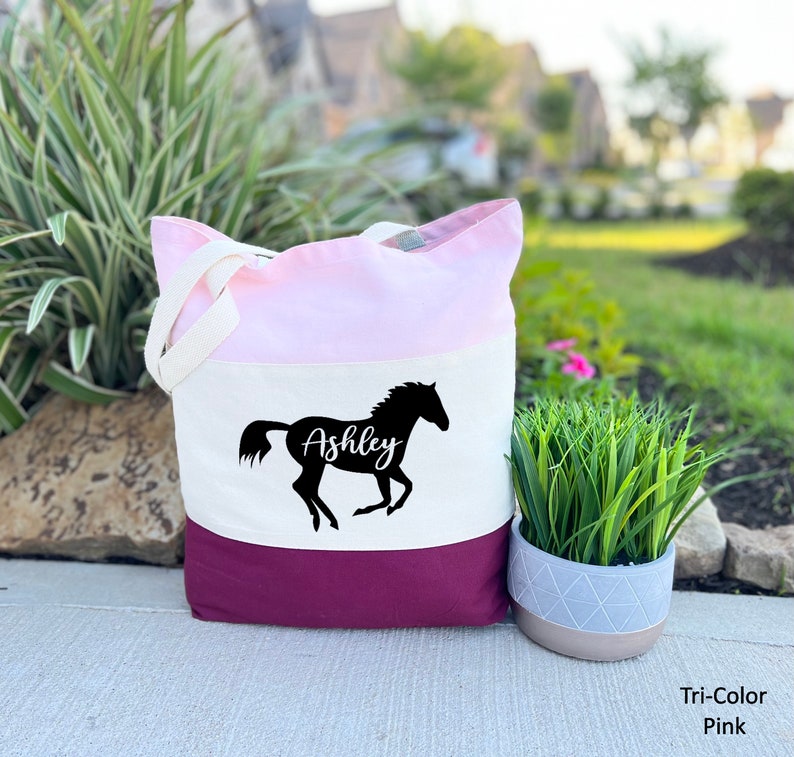 May include: A tri-color tote bag with a pink, white, and burgundy design. The bag features a black silhouette of a horse with the name "Ashley" written on it.