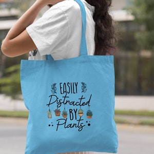 Pode incluir: Sacola de tecido azul claro com o texto "Easily Distracted By Plants" e um gr&aacute;fico de plantas em vasos pendurados no texto.