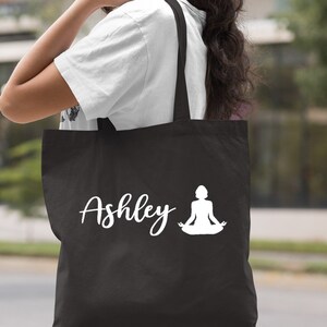 Pu&ograve; includere: Borsa tote in tela nera con testo bianco e una silhouette bianca di una persona in una posa di yoga. Il testo sulla borsa dice "Ashley".