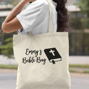May include: A natural canvas tote bag with black text that reads "Emmy's Bible Bag" and a black silhouette of a closed book with a cross on the cover.