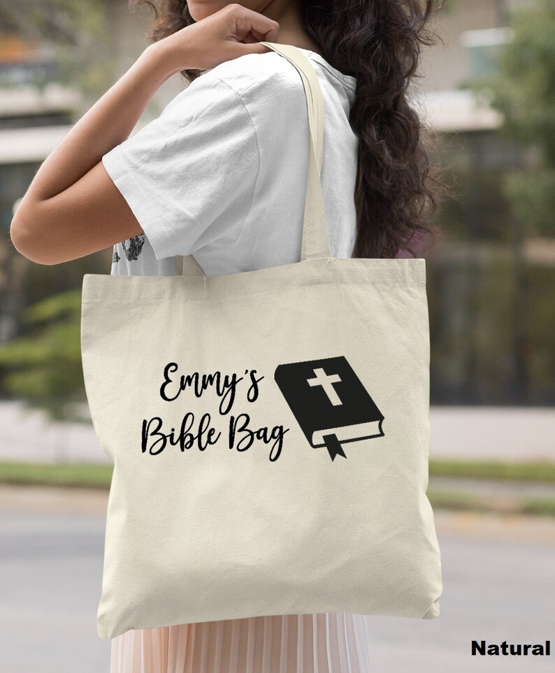 May include: A natural canvas tote bag with black text that reads "Emmy's Bible Bag" and a black silhouette of a closed book with a cross on the cover.